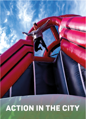 Eine Person springt von einer großen aufblasbaren Struktur vor einem strahlend blauen Himmel mit Wolken. Der Text am unteren Rand lautet: "ACTION IN THE CITY".
