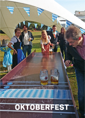 Menschen spielen auf einem Oktoberfest im Freien ein Bierglasschiebespiel, mit blauen und weißen Dekorationen und anderen Zuschauern in traditioneller bayerischer Kleidung. Das Wort OKTOBERFEST erscheint am unteren Rand.